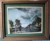 Framed artwork showing a mountain and creek scene with house, trees, and figures under cloudy sky, wood textured frame with green matting.