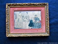 Close-up of one framed print showing three women and a child in historical dresses. Ornate gold-tone frame with pink matting.