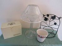 Photo showing frosted glass candle lamp, boxed Sam Seok mugs with lids, and metal flower pot hanger with flower pot, displayed on teal surface.