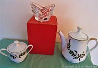 Porcelain tea pot and sugar bowl with holly design and gold trim beside Mikasa clear glass heart-shaped candy dish sitting on a red box
