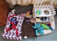Photo showing two dog beds, wooden dog bowl holder with ceramic bowls, various dog toys, two leashes, brown pet blanket, and bone-shaped pad.