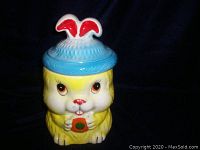 Front view of ceramic rabbit cookie jar with lid on showing yellow body, blue top with red ears.