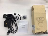 First black Full HD webcam with lens, USB cable, user manual, multifunction bracket, small accessory bag, and original cardboard box with webcam illustration.