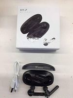 Top-down shot of the XY-7 packaging box alongside the open charging case showing earbuds inside and a white USB charging cable.