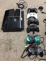 Full view of PlayStation 3 console and 6 controllers arranged on carpet including 4 black PS3 controllers, 1 white Xbox-style controller, 1 green wired controller.