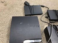 Overview photo showing PlayStation 3 slim console, PlayStation 2 original console, two PS2 DVD players with attached cables and power cords, all placed on carpeted floor.