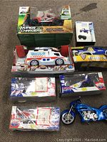 Top-down photo showing boxed 2014 Ford F150 Hyper Chargers truck, Hover Way micro drone, ambulance toy, Tonka Rescue Force truck, two Lucas Oil Racing cars, and blue display motorcycle model placed on carpet.