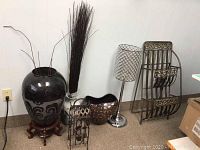 Photo showing 3 vases (black ceramic on wooden stand, tall white with sticks, bronze lattice), crystal-patterned chrome floor lamp, and metal ornate newspaper stand.