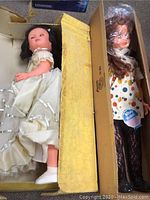 Two large porcelain dolls laying in cardboard, one in a white dress with satin trim, the other in a polka dot dress with curly hair