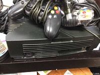 Two black original Xbox consoles shown with controllers and cables on top.