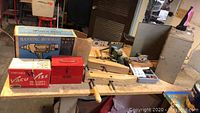 Wide shot showing lot contents including drill in box, clamps, vise, stapler, red toolbox, and hardware organizer on wooden bench