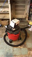 Craftsman red and black shop vacuum with attached hose sitting on tiled floor near door.