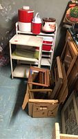 Photo of the drink cooler jug, drink thermos, crockpot, covered enamel dish and empty frames stacked beside a shelving unit.