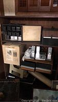 Overview of shelf with Bell & Howell projector box, slide projector, black slide cubes, AT&T cordless phones, filing supplies and storage boxes.