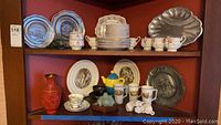 Display cabinet with multiple shelves holding Royal Albert china plates and teacups, pewter Historical series plates with embossed designs, Harkerware painted dishes, tall mugs including a yellow and blue spouted one, white cat-shaped salt and pepper shakers, and other ceramic items.