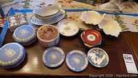 Photo showing an assortment of Wedgwood blue jasperware plates and lidded boxes, floral decorative plates, small bowls, a gravy boat, marble covered dish, and red lacquered box arranged on a table.