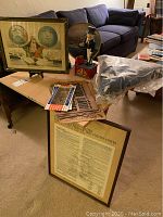 Photo showing a collection of Americana items on table with framed political cartoon, Uncle Sam figurine bank, Confederate currency, wanted posters, and a framed US Constitution bicentennial print leaning on floor.