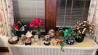 Photo of home decor items including watering cans, artificial plants, sea shells, and candles arranged on a surface in front of window blinds.
