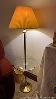Brass floor lamp with pleated beige fabric lampshade illuminated. The integrated glass side table is visible below the bulb.
