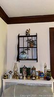 View of the wood wall shelf mounted on wall with various decorative items arranged on top and hanging plants inside shelf. Several items placed on surface below the shelf including glass oil lamp, steins, jars, candles etc.