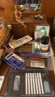 Overview of all items on a wooden table including mannequin, pastels boxes, lettering set, linoleum cutting tools, pencils in cup, eyeglasses, and other supplies.