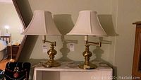 Pair of brass finish swing arm table lamps on a shelf, showing full height with white fabric shades.