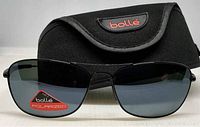 Front view of Bolle polarized aviator sunglasses with black metal frame and dark lenses with red Bolle polarized sticker on lens in front of black Bolle soft case.