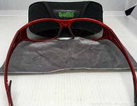Front view of red Bolle Turbulence sport sunglasses with black lenses, gray cleaning cloth laid out beneath, and black Bolle carrying case behind.