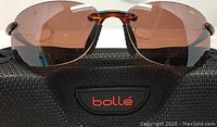Close-up of Bolle Kickback sunglasses with dark brown lenses and tortoise frame placed on the Bolle case showing rimless bottom lens and nose bridge detail.