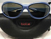 Front view of Bolle sunglasses with blue cobalt plastic frame and black polarized lenses displayed on black zippered case with red 'bollé' logo.