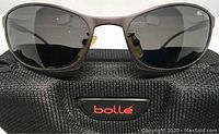 Front view of Bolle Montauk sunglasses with gray smoke polarized lenses and satin gun metal frame resting on Bolle case.