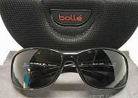 Front view of shiny black wrap sunglasses with gray polarized lenses, showing slight chip on left nose guard. Bolle branded case and cleaning cloth included.