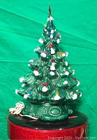 Front view of green glazed ceramic Christmas tree showing multicolored removable light bulbs, white snow accents, red star topper, decorative scroll-pattern base, and attached electric cord.