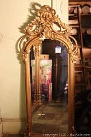 Full view of the large gilded mirror showing the tall rectangular shape and elaborate top carving, placed against a wall with visible floor and surrounding items.