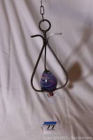 Full view of the oil lamp hanging in the wrought iron holder, showing the teardrop-shaped blue swirling glass reservoir and hanging chain.