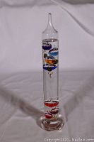 Full view of tall clear cylindrical Galileo thermometer with colorful floating bubbles inside liquid.