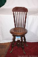 Full front view of vintage wooden piano stool chair with round seat and spindle backrest alongside the numbered tag.