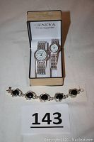 Geneva his and her watches with silver bands displayed in original box, alongside black and silver detailed bracelet.