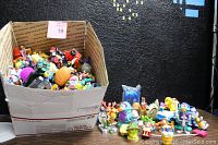 Wide shot showing a large open cardboard box filled with multiple small colorful PVC toys including action figures, cartoon characters, vehicles, and miscellaneous figures.