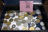 Photo showing a box containing multiple assorted antique and vintage coins and tokens from various countries with visible silver and bronze coins and tokens, including a large silver commemorative coin in protective casing and several smaller silver and brass coins.