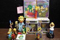 Assortment of Simpsons collectibles including DVDs, vinyl figures, plushie, PEZ dispenser, and multiple character figures.