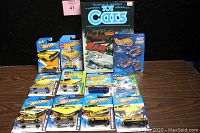 The full lot showing all 10 yellow Hot Wheels cars in packaging, 3 keyrings, and the toy cars collector's guide book, arranged on wooden surface with black background.