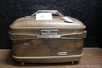 Exterior view of the brown American Tourister hard case luggage with combination lock and attached luggage tag, showing vintage sturdy design.