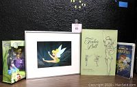 Group shot of all four items: framed Tinker Bell print, oversized Tinker Bell book, bath poufs in original packaging, and Peter Pan VHS tape.
