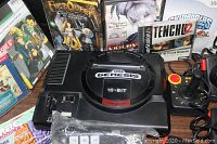 Photo of Sega Genesis 16-bit console with game cases in background and a joystick controller to the right.