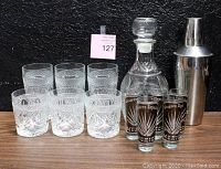 Photo showing six decorative glasses arranged behind three Reserva Especial Mexico shot glasses, glass decanter topped with stopper and text label, and stainless steel cocktail shaker on the right