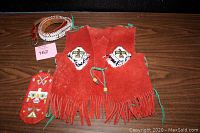 Photo showing the adult tooled leather beaded belt, child's suede beaded vest in rust color with beadwork, and leather beaded eyeglass case laid out on a table.