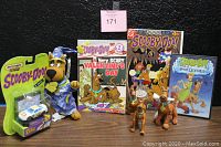 Nine Scooby-Doo themed items arranged on table including plush toy in wizard outfit, comics, DVDs, small figurines, and Matchbox car in original package.