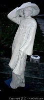Front angled view of white statue of woman in long dress and wide hat on black base, outdoor setting.