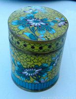 Oblique frontal view of the drum-shaped cloisonne box displaying yellow background with star motifs and floral designs in teal and pink.